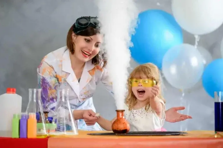 Science instructor in lab coat creating a dramatic science demonstration with white smoke while excited child in safety goggles watches with balloons in background